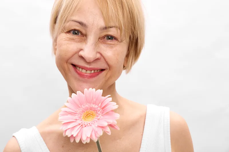 Senior woman smiling closeup portrait, holding pink gerbera flower near her cheek, soft studio light, white background, gentle mood, natural beauty