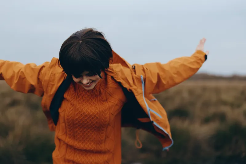 Smiling woman portrait in orange coat runs outdoors in open field with arms out, wind moves hair, cozy lifestyle mood in autumn under soft warm light on windy day