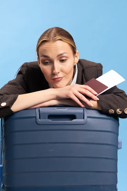 Tired woman resting on large blue suitcase holds passport and ticket, wearing brown jacket on light blue background. Concept of travel fatigue, waiting, and trip preparation.
