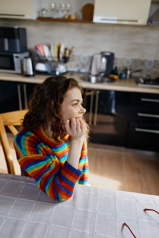 Thoughtful woman indoors, seated at a kitchen table, hands under chin, rainbow striped sweater, warm natural light, calm domestic moment, cozy home atmosphere, everyday lifestyle