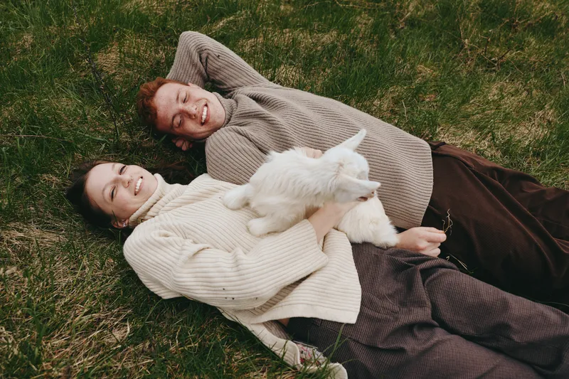Two young women lie on grass holding a white baby goat while smiling and enjoying a relaxed lifestyle moment outdoors in casual sweaters and pants.