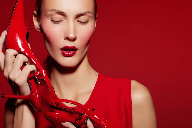 Close-up of a woman with red lipstick holding red high heels near her face against a red background. Concept of fashion, beauty, style, and elegance with vibrant colors.