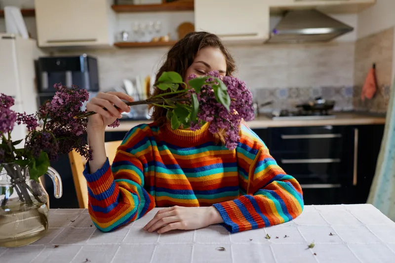 A young brunette woman wearing a colorful striped sweater sits at a dining table in a bright kitchen, smelling a lilac bouquet, her calm expression hinting at mindfulness.