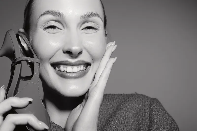 Smiling caucasian woman with fair skin holds a high heel shoe near her face, expressing joy and excitement. Studio portrait with soft lighting on plain background.