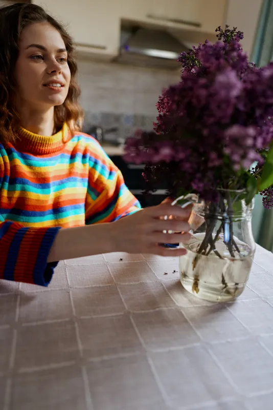 A woman in a bright striped sweater sits at a dining table inside a cozy home, gently arranging purple flowers in a vase, capturing everyday lifestyle, home decor, and creative stillness.