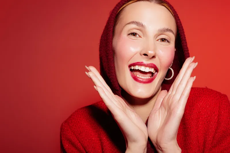 Happy woman in red hoodie smiles brightly with hands near face on red background. Concept of joy, fashion, and positive emotions in studio portrait photography.