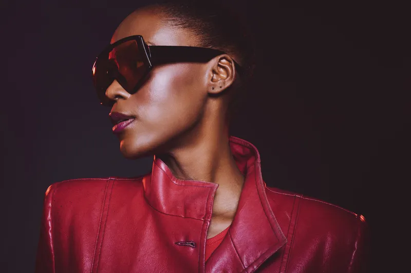 Stylish African woman with dark skin wearing oversized sunglasses and a red leather jacket poses confidently on black background. Concept for fashion, elegance, and modern style.