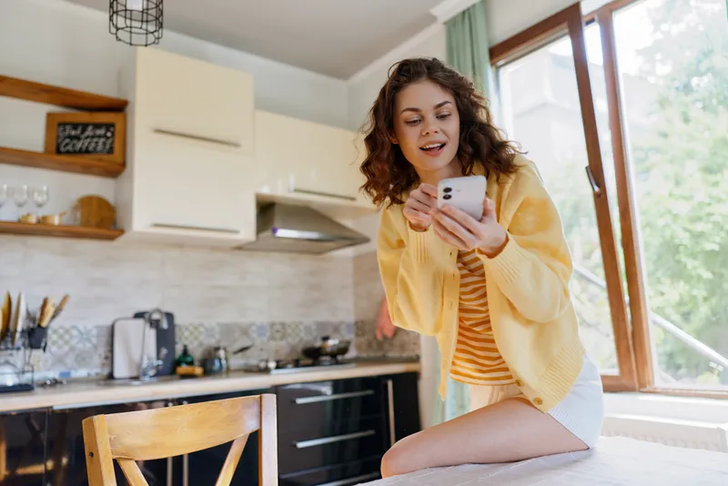 A woman in a sunny kitchen sits on a stool, scrolling on a smartphone, wearing a yellow cardigan and striped top. Casual home lifestyle, everyday tech use, candid moment.