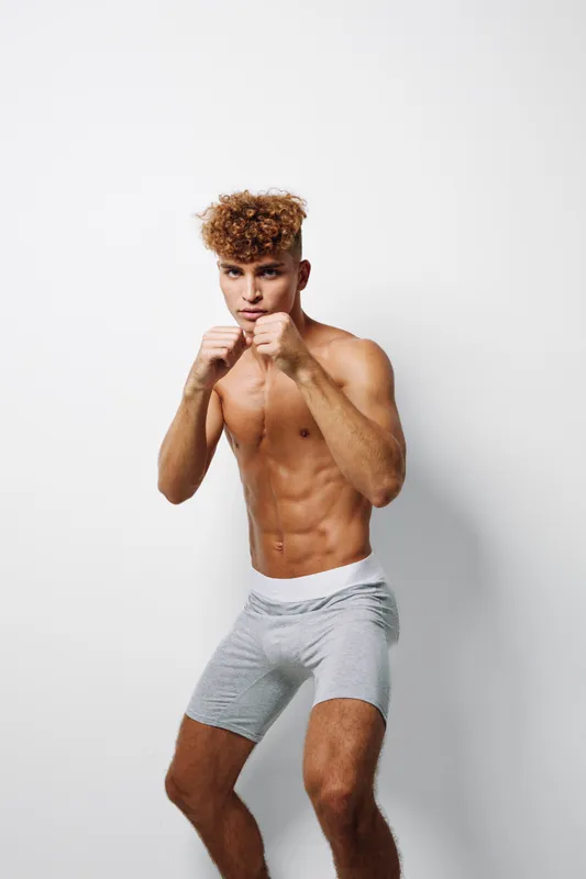 Young athletic man with curly hair in boxing stance wearing gray shorts against white background. Fitness, training, workout and strength concept with muscular body and focused expression.