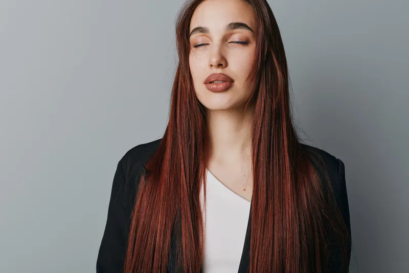 Young woman with long straight hair and closed eyes posing in studio. Female model wearing black jacket and white shirt with natural makeup on gray background for portrait.