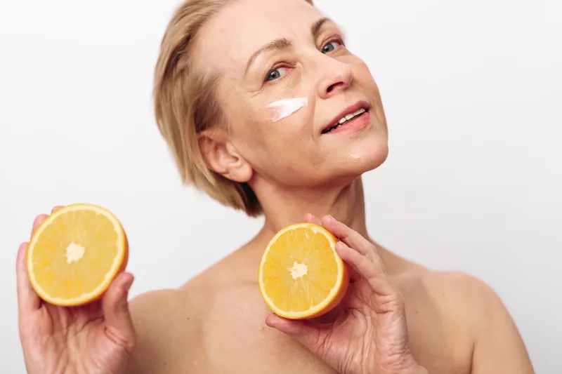 Mature woman with short hair holds two orange halves near her chest, smiling gently. Visible facial cream on cheek suggests skincare, natural beauty, health, and wellness concept in studio.