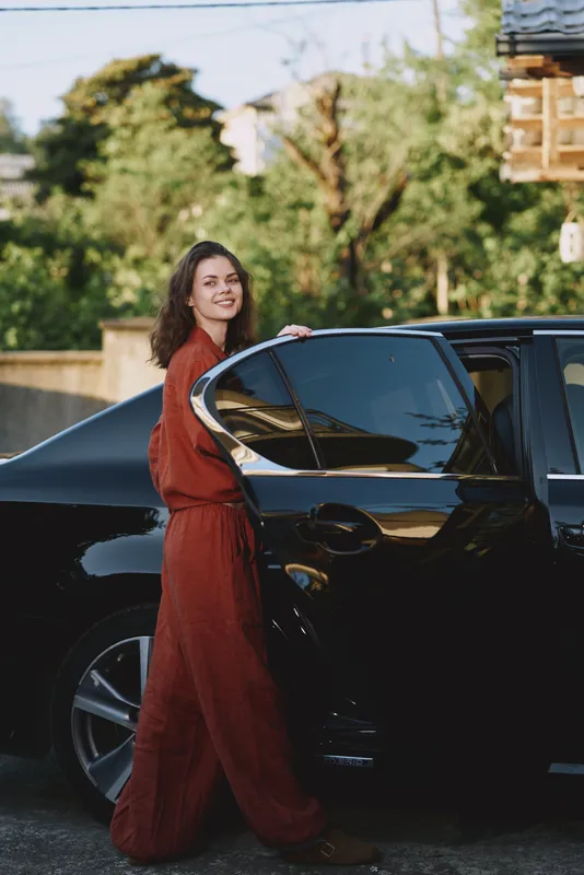 Young woman with long brown hair in a rust jumpsuit stands beside a glossy black car in a sunny driveway, smiling warmly, conveying a casual street vibe with natural light and a confident, relaxed