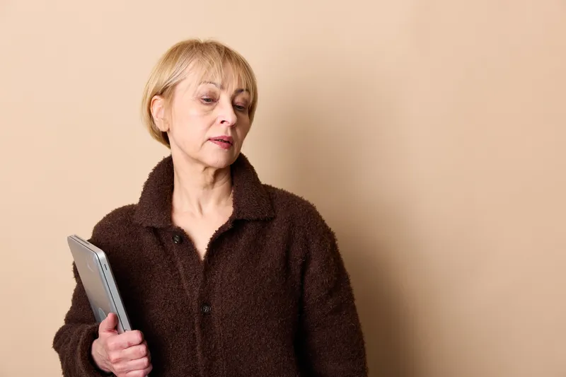 Elderly woman using tablet indoors, beige background, close up portrait, serious focused expression, digital technology for reading and online communication, home routine, copy space