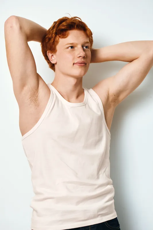 Young man with red hair and fair skin wearing white tank top posing relaxed with hands behind head against white background in studio lighting.