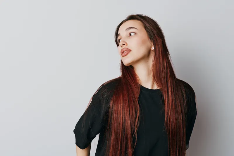 Young woman with long red hair wearing black shirt looking sideways on light background. Portrait of female model with natural makeup and straight hair in casual style indoors.