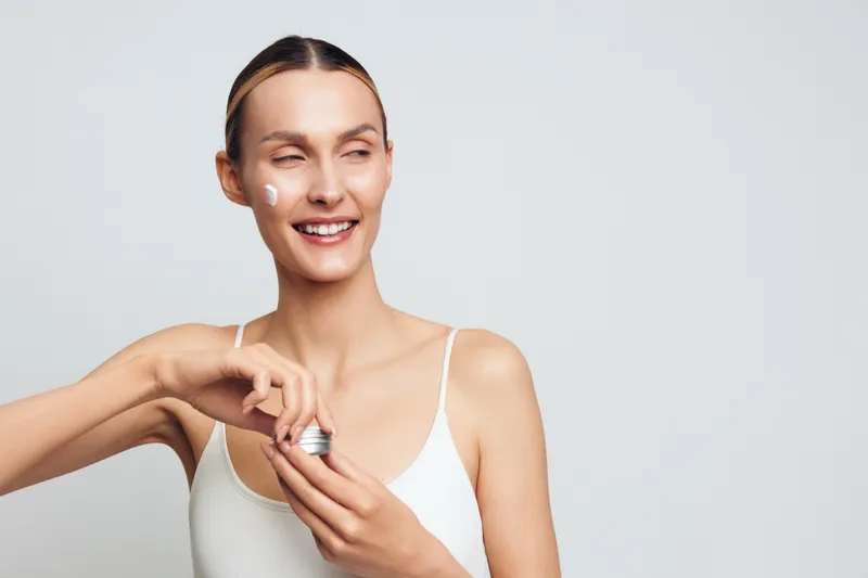 Smiling woman applies face cream in a studio close up with soft light on a light gray background holding a small applicator for self care beauty skincare wellness and fresh skin