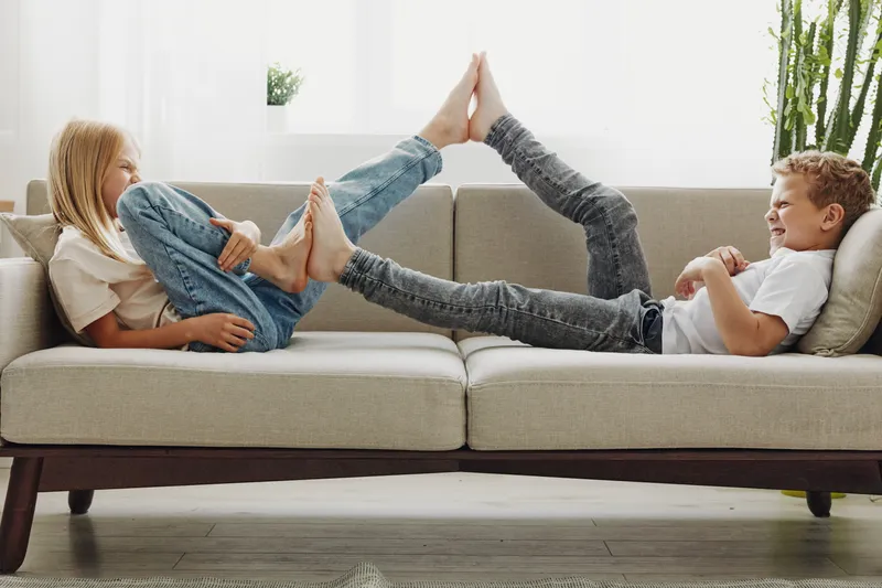 Happy children lying on sofa playfully touching feet, smiling and enjoying leisure time in bright living room. Concept of sibling bonding, childhood fun, and family relaxation moments.