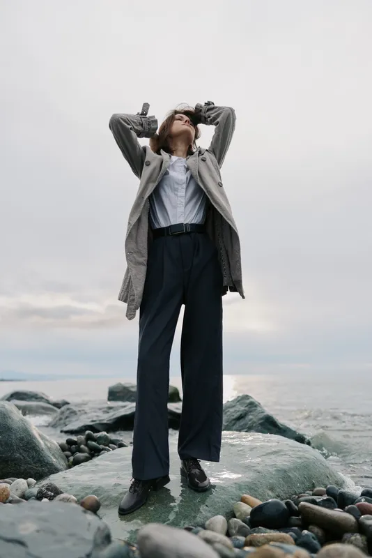 Tall person in stylish outfit stands on rocky shore with hands raised behind head, looking up to cloudy sky. Fashion concept with moody natural landscape and casual modern clothing.