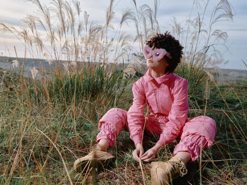 Outdoor girl in a pink outfit sits among tall grasses, wearing a playful mask, a calm reflective moment in nature amid a wide open landscape.