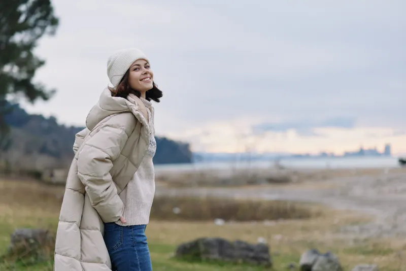 Winter woman in a puffy coat, scarf and beanie outdoors along a shoreline, smiling softly as the chilly breeze and expansive landscape frame a calm, reflective moment.