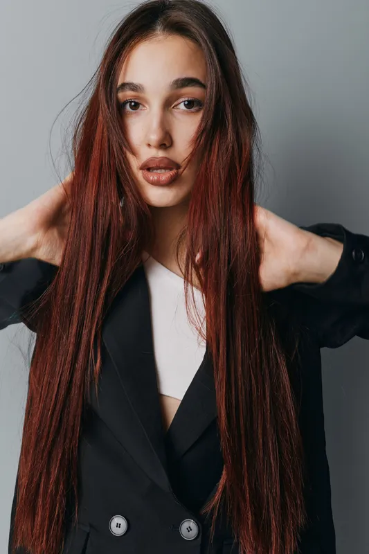Young woman with long straight hair wearing black blazer and white top posing with hands behind head against gray background in studio portrait fashion style beauty.