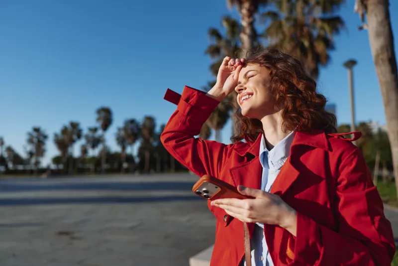 Young woman in red coat smiles holding smartphone outdoors under clear blue sky with palm trees in background enjoying sunny day and fresh air in urban park.