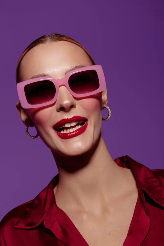 Stylish woman with pink sunglasses and red lipstick smiles confidently against purple background. Fashion portrait with bold makeup and trendy accessories in studio lighting.