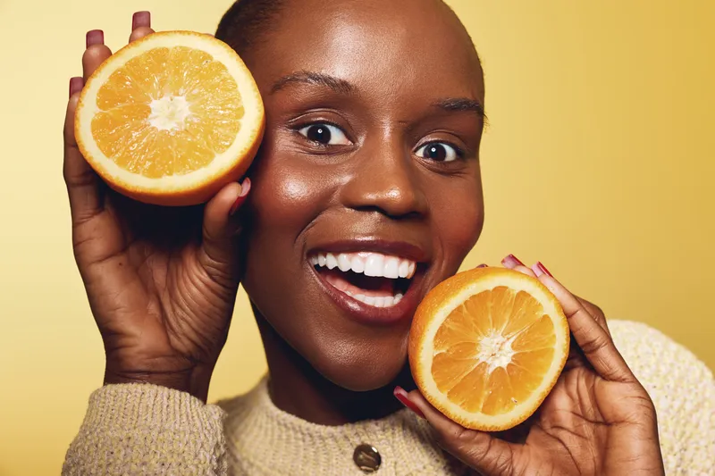 Happy African woman with dark skin holding two orange halves near face on yellow background. Concept of fresh fruit, healthy lifestyle, wellness, and natural beauty close-up portrait.