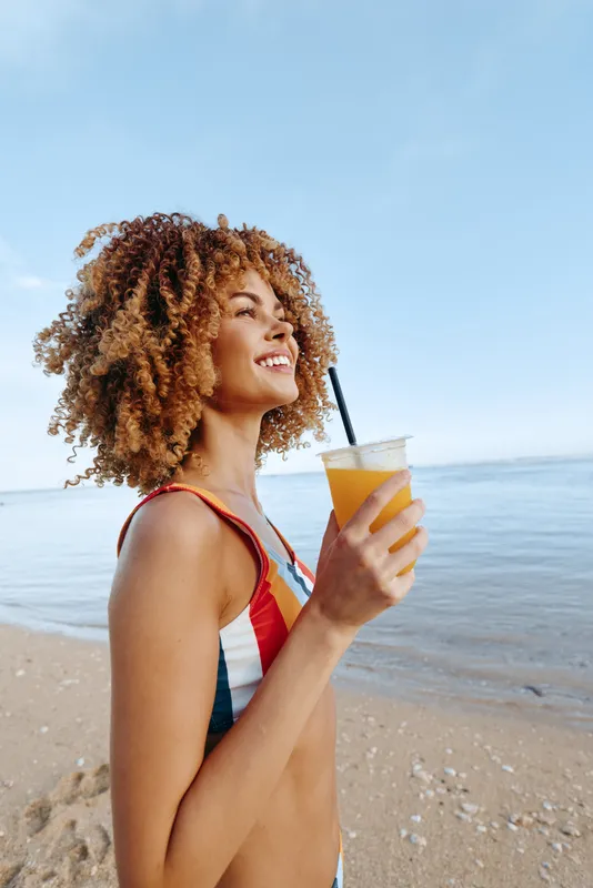 Woman with curly hair on the beach, holding a drink of orange juice, relaxing in summer sunlight, ocean horizon behind, seaside vacation mood, natural daylight