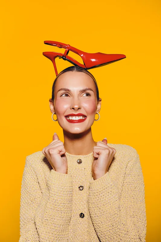 Smiling woman with red high heel shoes on her head poses against bright yellow background. Concept of fashion, fun, creativity, and playful style with vibrant colors.