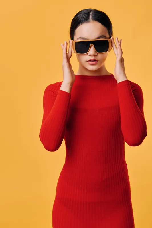 Woman in red dress adjusting black sunglasses standing against yellow background. Fashion model with serious expression wearing slim fit knitted long sleeve outfit in studio.