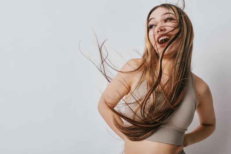 Woman with long hair smiling and posing against white background indoors. Casual style, happy expression, energetic movement with loose hair in studio lighting.