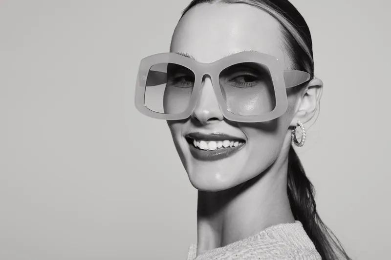 Smiling white woman with fair skin wearing large glasses and earrings looks at camera. Studio portrait in black and white with clean background.