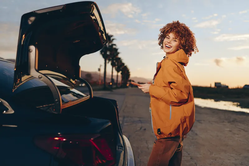 Woman lifestyle car jacket sunset outdoors road travel orange curly hair smile beside open door, outdoors travel mood in golden evening light, casual portrait concept, road trip by the coast