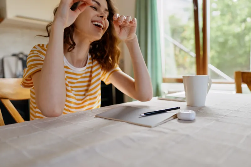 Young woman with wavy hair sits at a sunlit kitchen table, smiling as she writes on a sheet, with a mug nearby, creating a warm, candid home workspace moment.