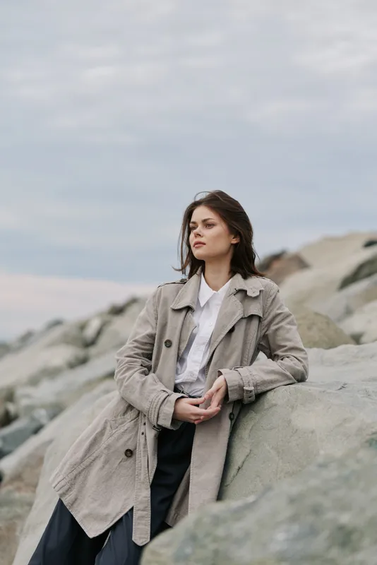 Young woman in beige coat leans on large rocks outdoors under cloudy sky. She looks away thoughtfully, wearing casual pants and white shirt. Concept of autumn fashion and natural portrait.