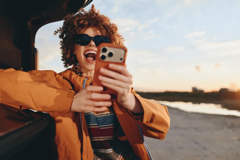 Woman lifestyle selfie, holding smartphone with sunglasses and curly hair, smiling outdoors by the seaside at sunset, wearing an orange jacket, candid cheerful coastal portrait