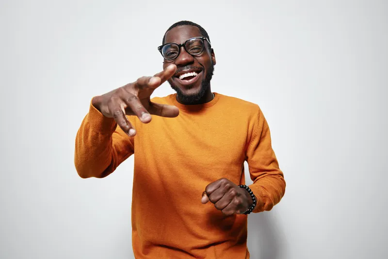 African American man with dark skin wearing glasses and orange sweater smiling and pointing at camera in studio. Happy male portrait with casual clothing and positive expression on white background.