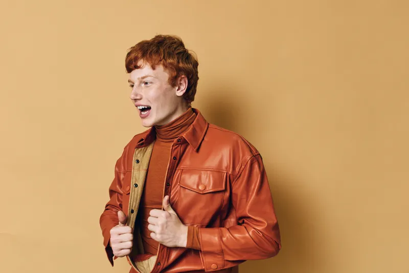 Young man with red hair wearing a brown turtleneck and orange leather jacket posing smiling with open mouth against beige background in studio. Casual fashion style portrait.