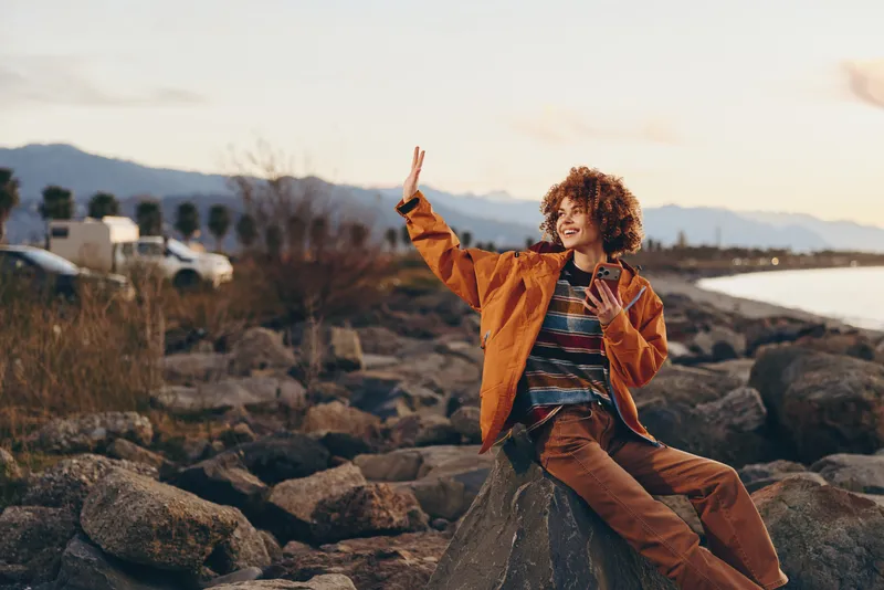 Woman lifestyle outdoors seaside rocks at sunset, curly haired woman smiling, outstretched arm pose, orange jacket striped shirt, calm sea and distant mountains, golden light, travel mood