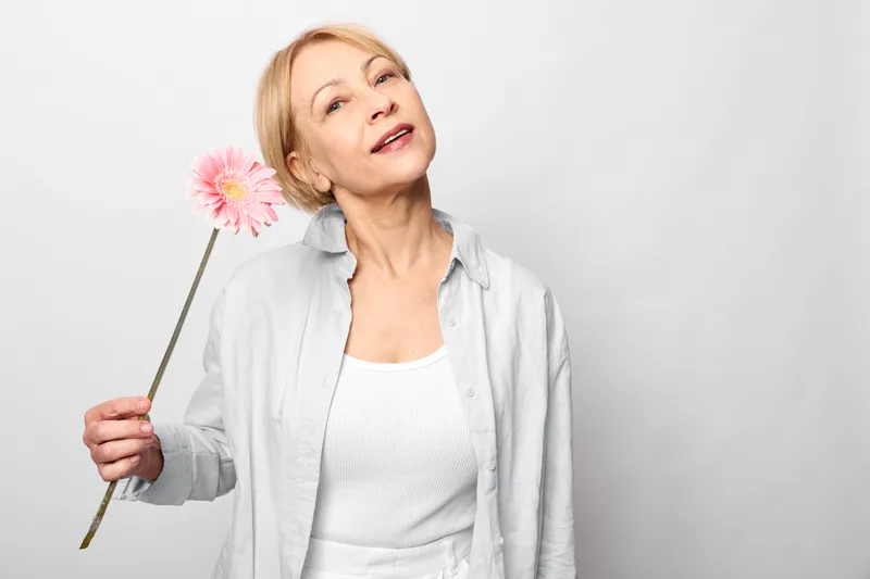 Elderly woman holding pink daisy in studio, she shows sneeze reaction with watery eyes, stressed expression, health concept allergy discomfort seasonal spring