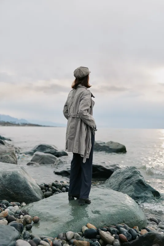 Woman in grey coat and hat stands on rocky shore looking at calm sea under cloudy sky. Concept for solitude, contemplation, nature, and peaceful outdoor moments by water.
