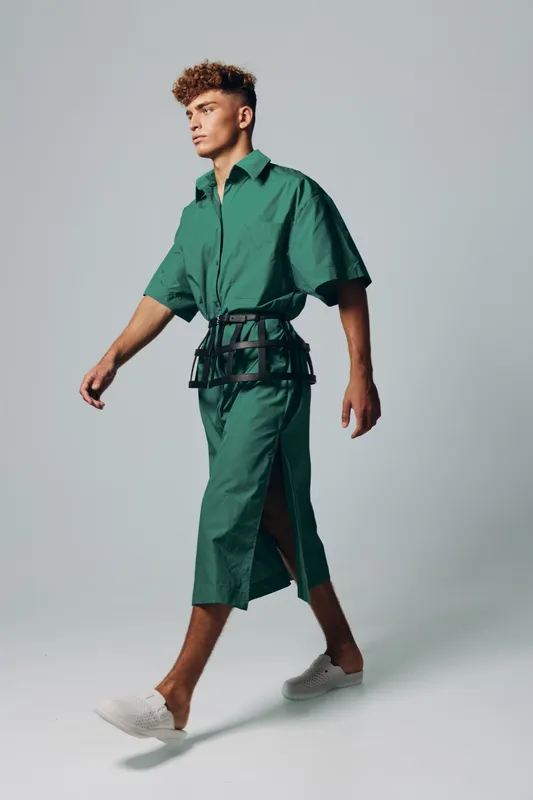 Young man walking in green oversized shirt and matching wide pants with white sneakers on plain background. Fashion model with curly hair in casual modern outfit.