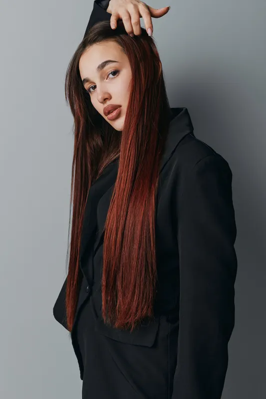 Young woman with long braided hair wearing black blazer posing confidently against gray background. Fashion portrait with stylish look and natural makeup for beauty and style concept.