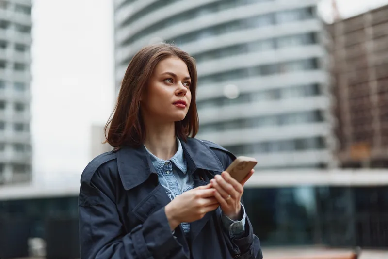 A young adult woman with brown hair walks in a city, looking at her smartphone, wearing a denim jacket over a light blue shirt, focused and thoughtful in a bustling urban environment