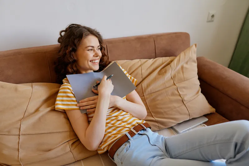 Woman relaxing on a couch at home, smiling while writing in a notebook, captured in a cozy living room scene that highlights casual daily life and personal moments for lifestyle use