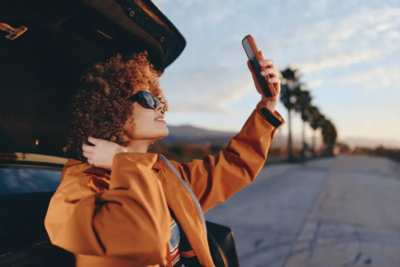 Woman with curly hair and sunglasses in a car at golden hour, taking a selfie with a smartphone. Orange jacket, outdoor city road background, travel lifestyle, warm mood today.