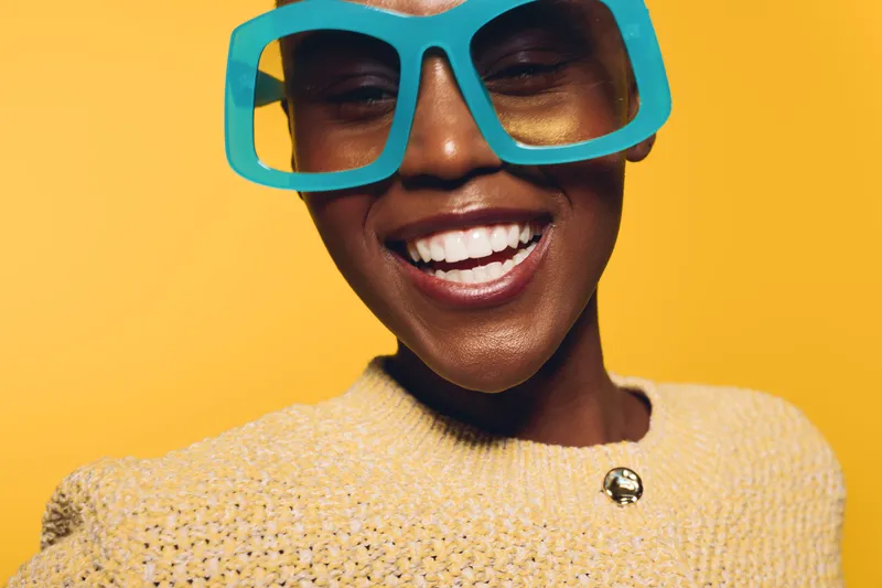 Close-up of smiling African American woman with dark skin wearing large blue glasses and yellow sweater. Bright yellow background creates cheerful and vibrant mood. Concept of happiness and fashion.
