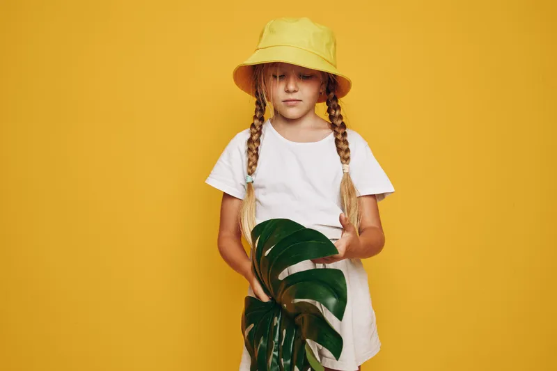 Young girl with braided hair wearing a yellow hat and white dress holding green tropical leaves on a yellow background.