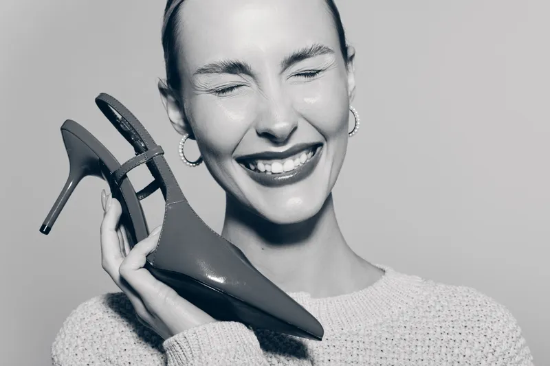 Smiling Caucasian woman with light skin holds red high heel shoe close to face, eyes closed, wearing white sweater and hoop earrings in soft studio lighting. Concept of fashion.